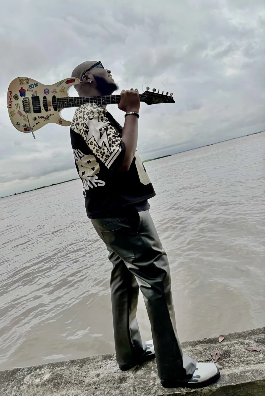 CJ Obassey holding his electric guitar across his shoulders by the water