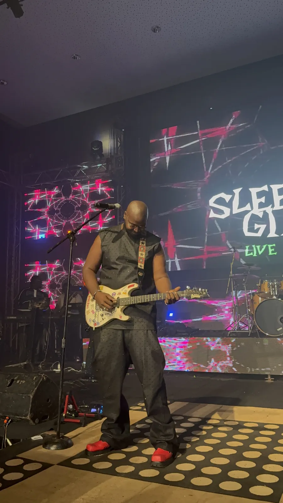 CJ Obassey playing his white electric guitar on stage, Sleeping Giant on the screen behind him