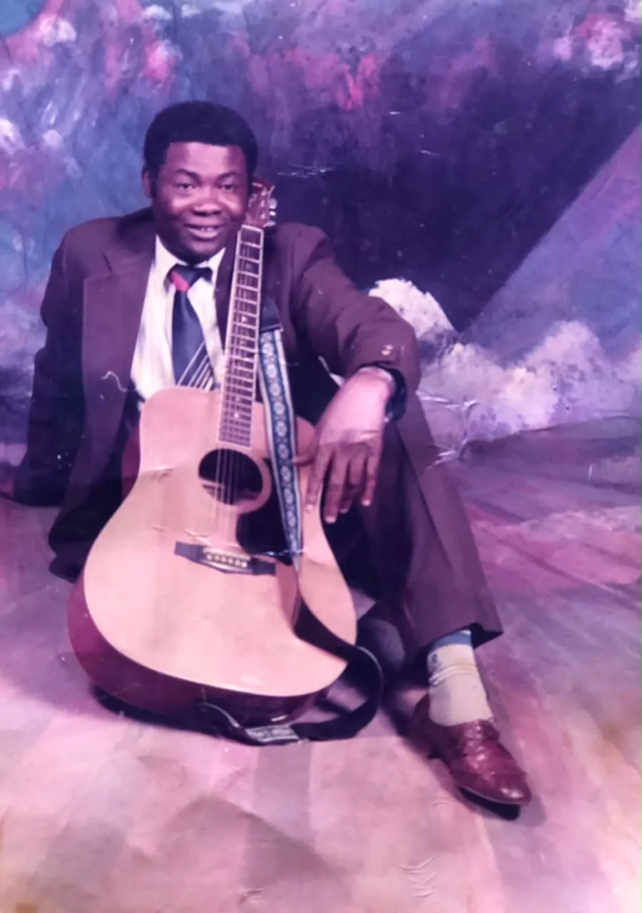 Rev. Patty Obasi seated with his acoustic guitar in a studio portrait